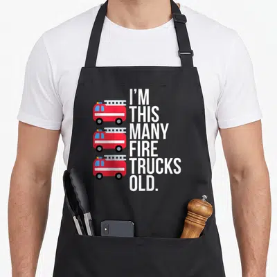 Fire birthday truck apron - Three stacked red fire trucks next to vertical white text reading I'm This Many Fire Trucks — shown on a black cotton canvas apron against a white background