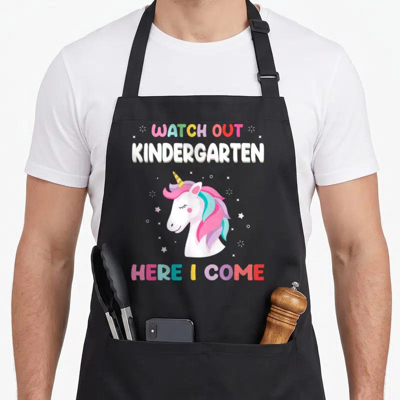 Watch kindergarten unicorn apron - Cartoon unicorn head with WATCH OUT KINDERGARTEN HERE I COME text and scattered — shown on a black cotton canvas apron against a white background