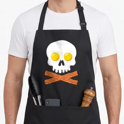 Breakfast bacon apron - Cartoon white skull with yellow egg yolk eyes and two crossed orange bacon strips below — shown on a black cotton canvas apron against a white background