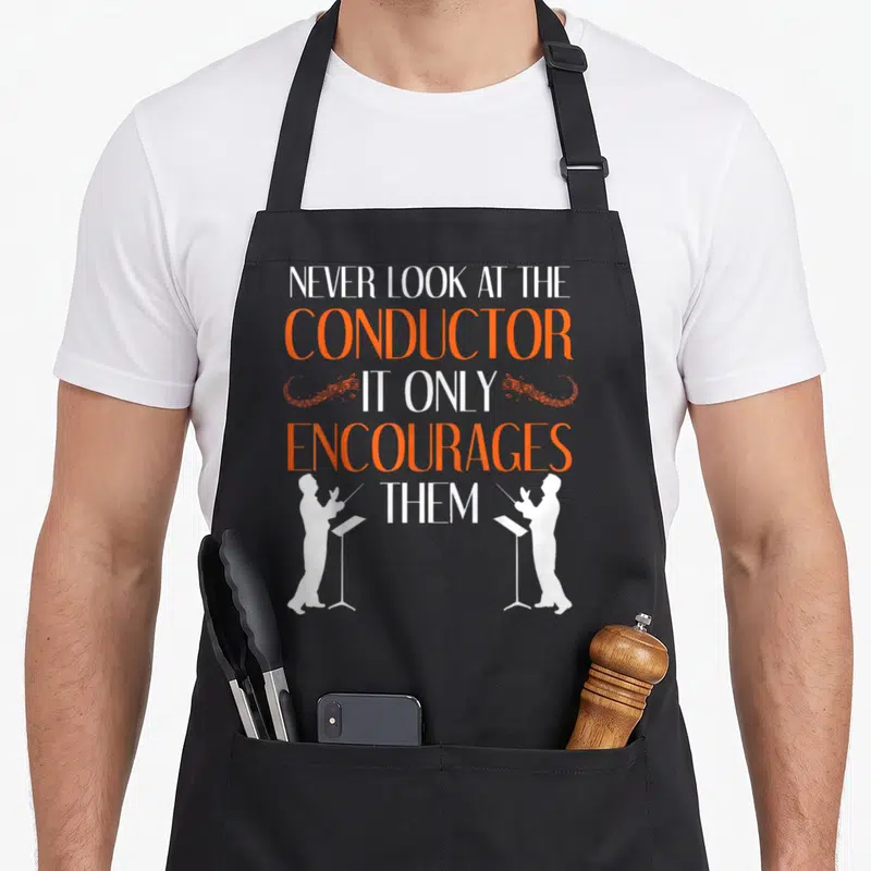 Conductor symphony life apron - Stacked white and orange text reading NEVER LOOK AT THE CONDUCTOR IT ONLY ENCOURAGES — shown on a black cotton canvas apron against a white background