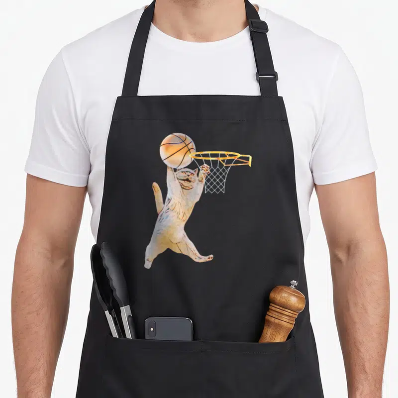 Basketball cat apron - Cartoon white cat jumping to shoot orange basketball into hoop with yellow rim — shown on a black cotton canvas apron against a white background