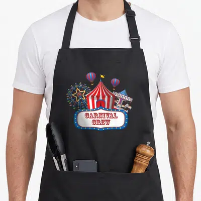 Carnival crew birthday apron - Circus tent, ferris wheel, carousel, two balloons, and CARNIVAL CREW text on a blue sign — shown on a black cotton canvas apron against a white background