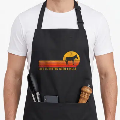 Better mule apron - Mule silhouette over yellow sun and orange red stripes with LIFE IS BETTER WITH A MULE text — shown on a black cotton canvas apron against a white background