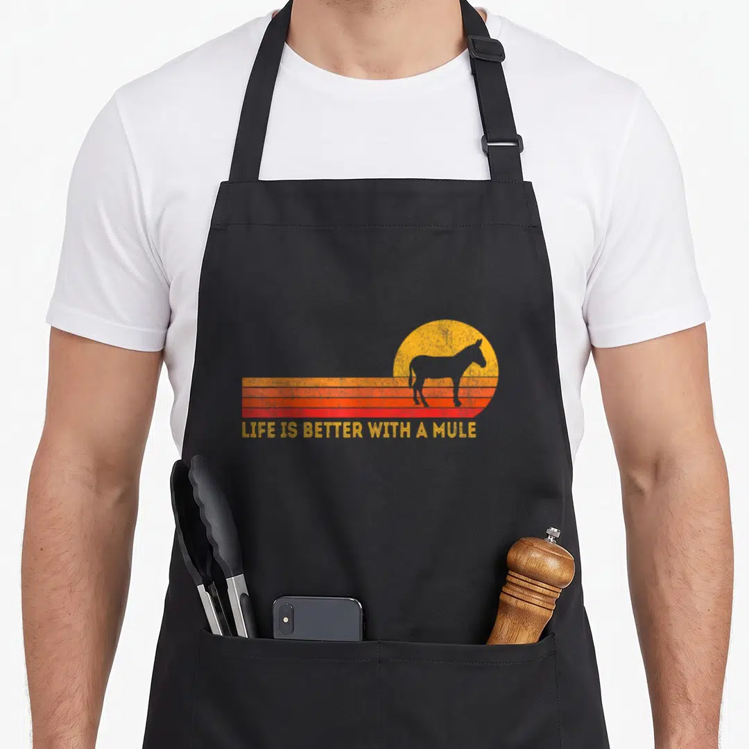 Better mule apron - Mule silhouette over yellow sun and orange red stripes with LIFE IS BETTER WITH A MULE text — shown on a black cotton canvas apron against a white background