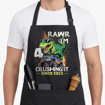 4th birthday monster truck apron - T-Rex in sunglasses and party hat on blue monster truck with celebratory text — shown on a black cotton canvas apron against a white background