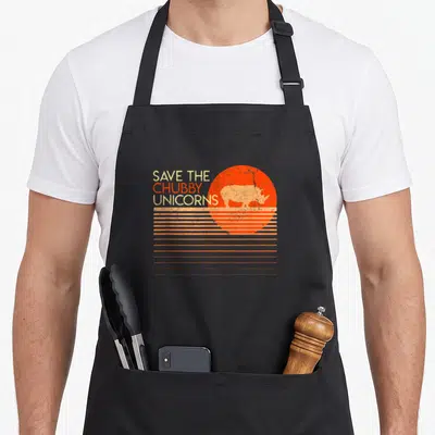 Save chubby unicorn apron - Retro rhino silhouette in orange sun with SAVE THE CHUBBY UNICORNS text — shown on a black cotton canvas apron against a white background