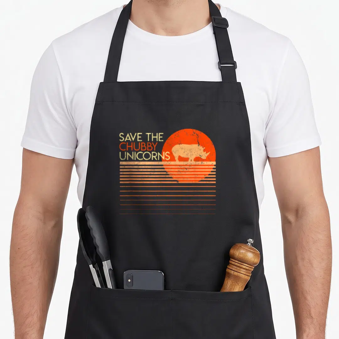 Save chubby unicorn apron - Retro rhino silhouette in orange sun with SAVE THE CHUBBY UNICORNS text — shown on a black cotton canvas apron against a white background