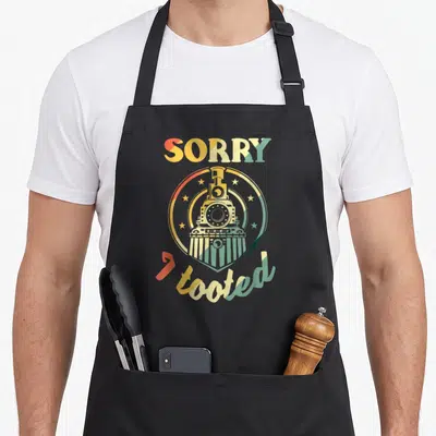 Tooted i love trailer apron - Retro steam train emblem with SORRY I tooted text in red yellow green and white — shown on a black cotton canvas apron against a white background