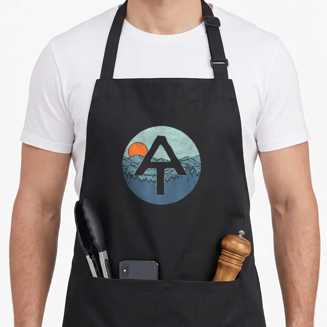 Appalachian outdoor apron - Grey letter A over circular blue mountain and orange sun landscape in retro style — shown on a black cotton canvas apron against a white background