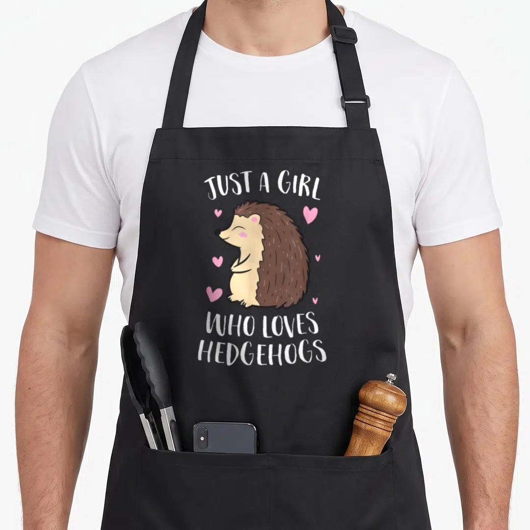 Cute hedgehog mom apron - Cartoon brown hedgehog with pink hearts and white text JUST A GIRL WHO LOVES HEDGEHOGS — shown on a black cotton canvas apron against a white background
