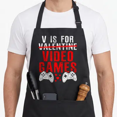 Video game valentine's day apron - V IS FOR VALENTINE crossed out with VIDEO GAMES text above two white controllers — shown on a black cotton canvas apron against a white background