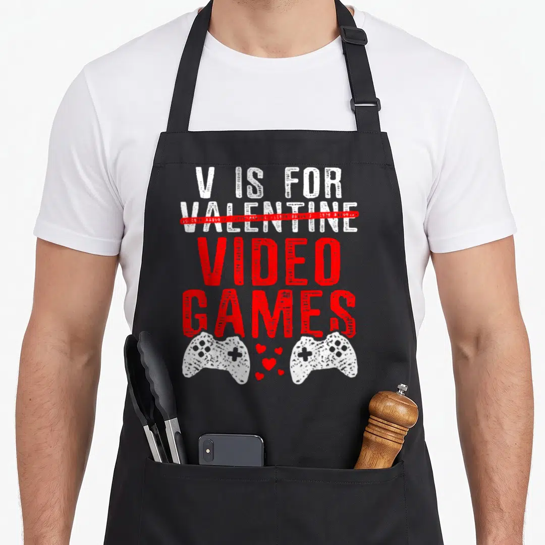 Video game valentine's day apron - V IS FOR VALENTINE crossed out with VIDEO GAMES text above two white controllers — shown on a black cotton canvas apron against a white background