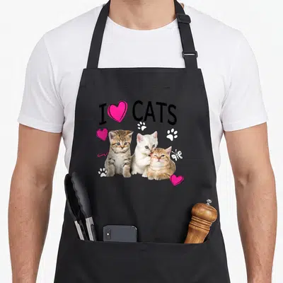 Kitten paw cat apron - I Heart Cats text above three kittens with pink hearts and white paw prints — shown on a black cotton canvas apron against a white background