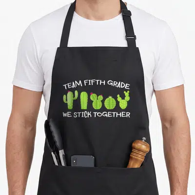Stick teacher apron - Five cartoon cacti with pink flowers and white text reading TEAM FIFTH GRADE WE STICK TOGETHER — shown on a black cotton canvas apron against a white background