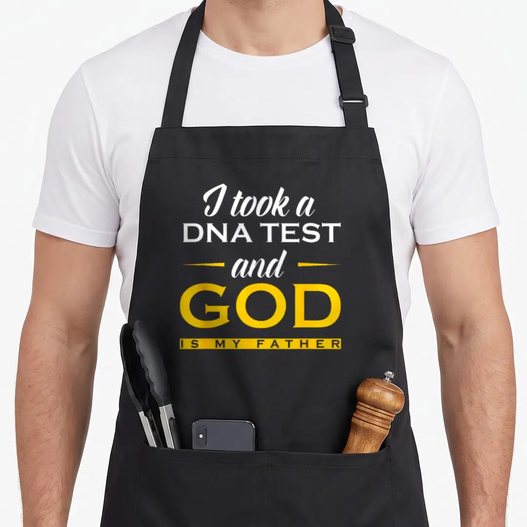 Took test kitchen apron - Stacked white and yellow text reading I took a DNA TEST and GOD IS MY FATHER with a yellow — shown on a black cotton canvas apron against a white background