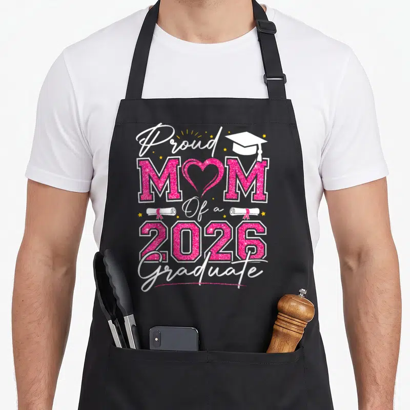 Proud graduation vibes apron - Stacked pink glitter text Proud MOM Of a 2026 Graduate with heart O, cap, and diplomas — shown on a black cotton canvas apron against a white background