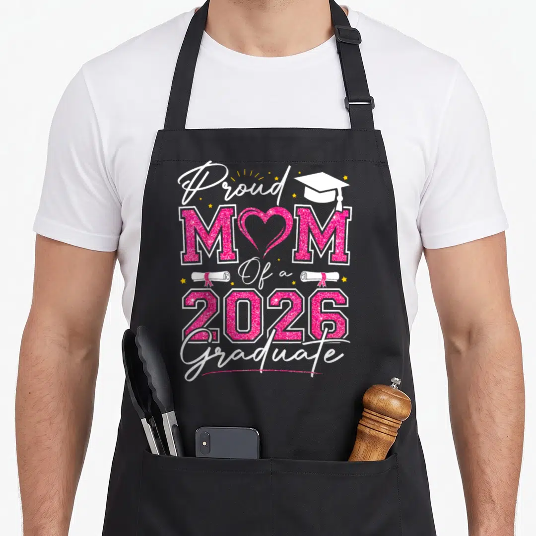 Proud graduation vibes apron - Stacked pink glitter text Proud MOM Of a 2026 Graduate with heart O, cap, and diplomas — shown on a black cotton canvas apron against a white background