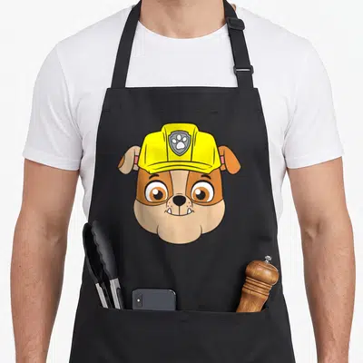 Rubble face apron - Cartoon bulldog head in yellow hat with paw print emblem, tan and brown colors — shown on a black cotton canvas apron against a white background