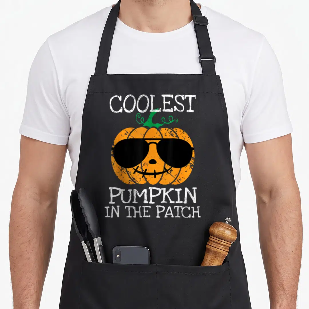 Coolest halloween apron - Orange pumpkin wearing black sunglasses with white text COOLEST PUMPKIN IN THE PATCH — shown on a black cotton canvas apron against a white background