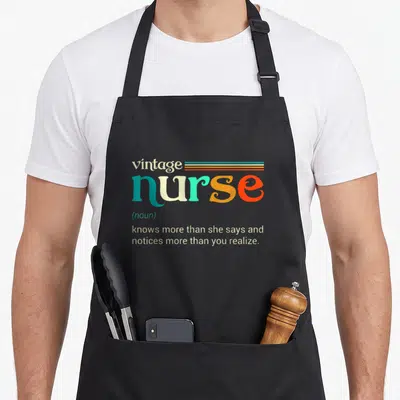 Noun nurse apron - Retro typography defining vintage nurse as knowing more than she says and noticing more than you — shown on a black cotton canvas apron against a white background