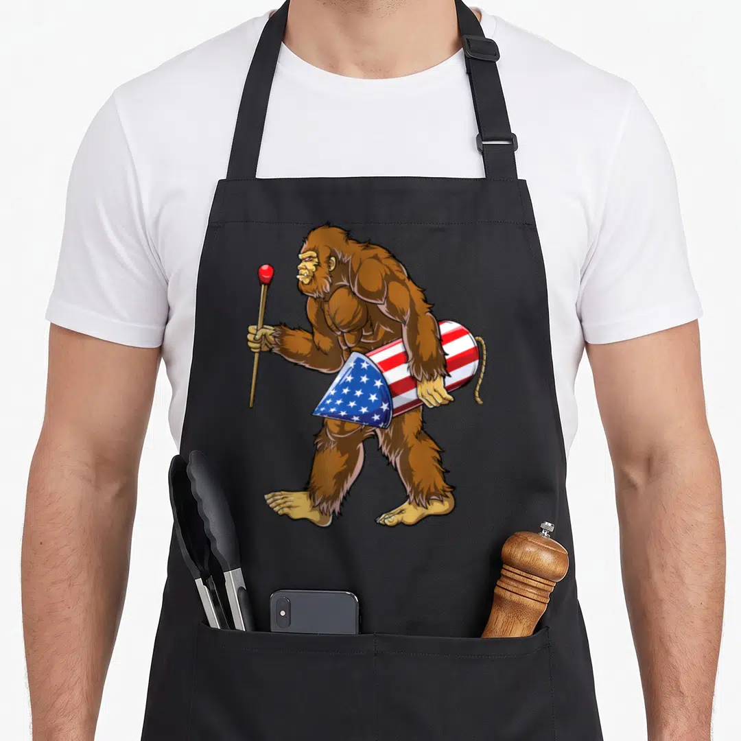 Sasquatch 4th of july apron - Cartoon brown bigfoot facing left holding staff and carrying red white blue star-spangled — shown on a black cotton canvas apron against a white background