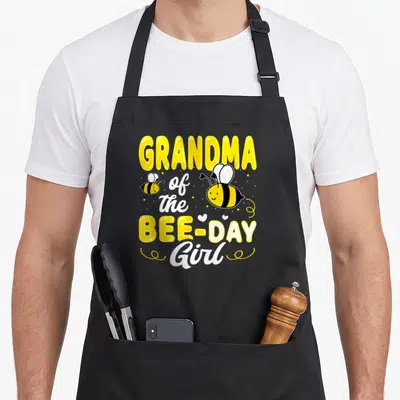 Grandma bee aprons - Stacked text GRANDMA of the BEE-DAY Girl with two cartoon bees and hearts — shown on a black cotton canvas apron against a white background