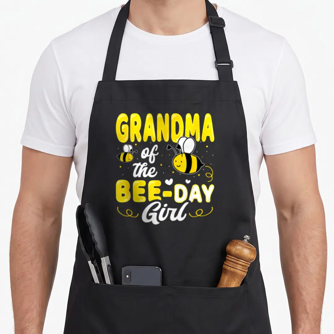 Grandma bee aprons - Stacked text GRANDMA of the BEE-DAY Girl with two cartoon bees and hearts — shown on a black cotton canvas apron against a white background