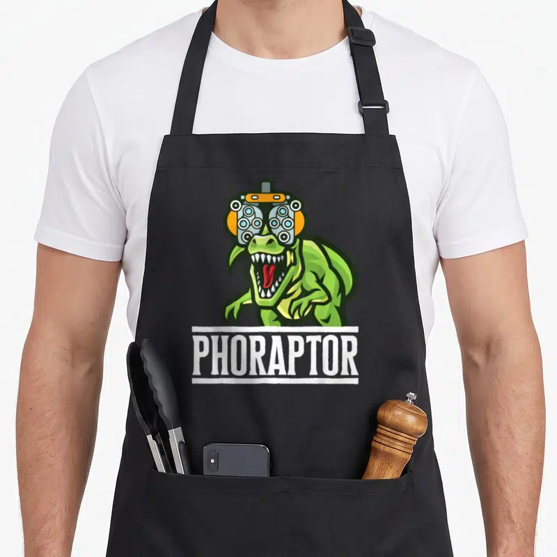 Optometry machine apron - Green dinosaur wearing a phoropter with white PHORAPTOR text banner below — shown on a black cotton canvas apron against a white background
