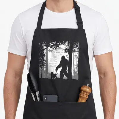 Sasquatch wood apron - Grey and white silhouette of Bigfoot walking a dog with UFOs in a forest — shown on a black cotton canvas apron against a white background