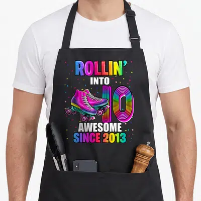 Skating 10th birthday apron - Rollin into 10 Awesome since 2013 roller skates rainbow text confetti — shown on a black cotton canvas apron against a white background