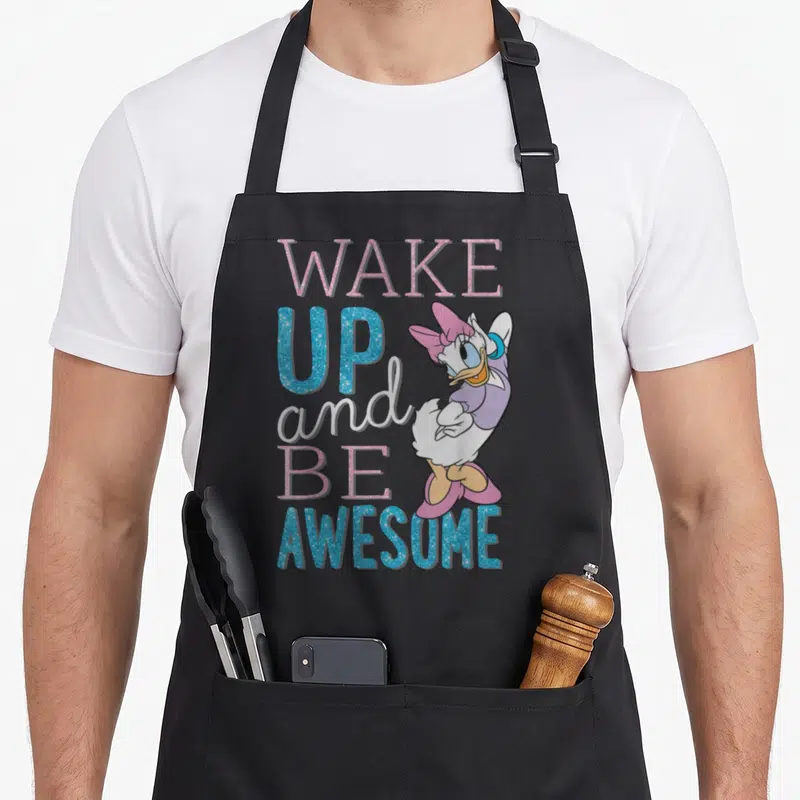 Cute daisy apron - Cartoon duck standing next to stacked WAKE UP and BE AWESOME text in pink, blue, white, and purple — shown on a black cotton canvas apron against a white background