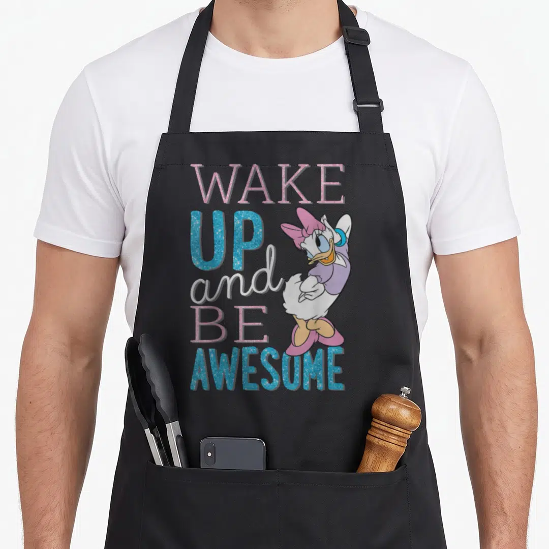 Cute daisy apron - Cartoon duck standing next to stacked WAKE UP and BE AWESOME text in pink, blue, white, and purple — shown on a black cotton canvas apron against a white background