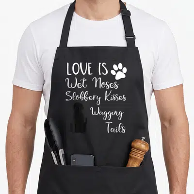 Dog labrador apron - White LOVE IS text and sitting black dog illustration — shown on a black cotton canvas apron against a white background