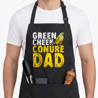 Cheek conure bird apron - Stacked Green Cheek Conure Dad text with cartoon parrot in green yellow and red — shown on a black cotton canvas apron against a white background