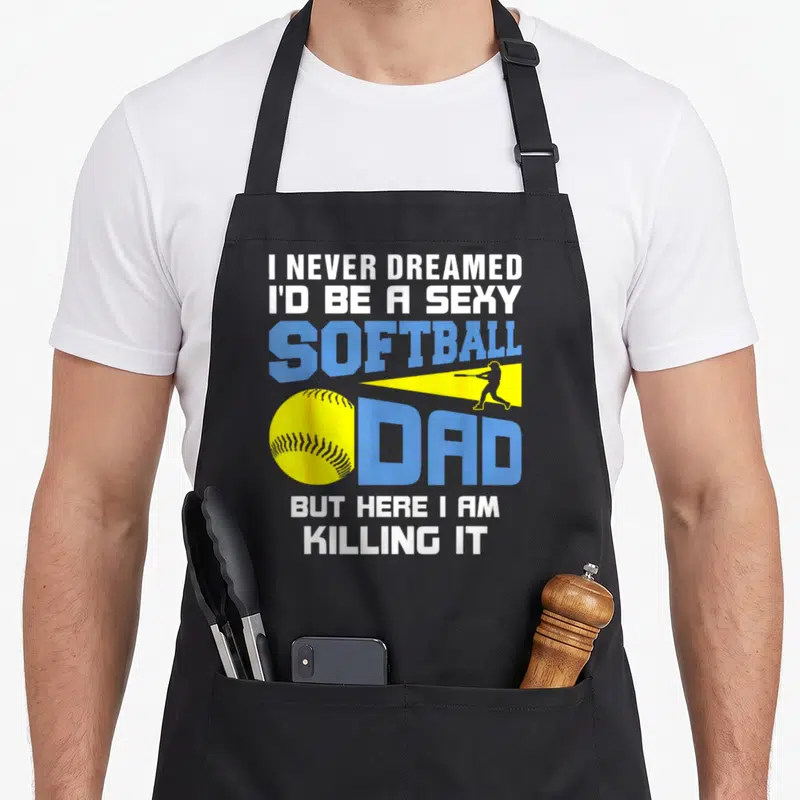 Dreamed i love watching apron - Stacked text I Never Dreamed Sexy Softball Dad with yellow ball and blue batter — shown on a black cotton canvas apron against a white background
