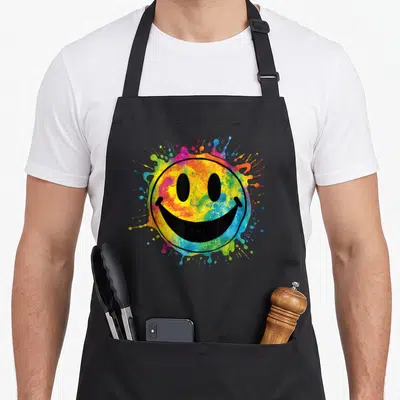 Rainbow face on apron - Black outlined smiley face over multicolored paint splatters in yellow, blue, red, and green — shown on a black cotton canvas apron against a white background
