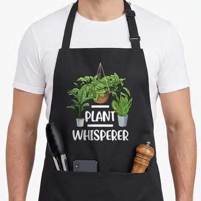 Whisperer gardener apron - Three houseplants including a hanging vine above PLANT WHISPERER text in white cartoon — shown on a black cotton canvas apron against a white background