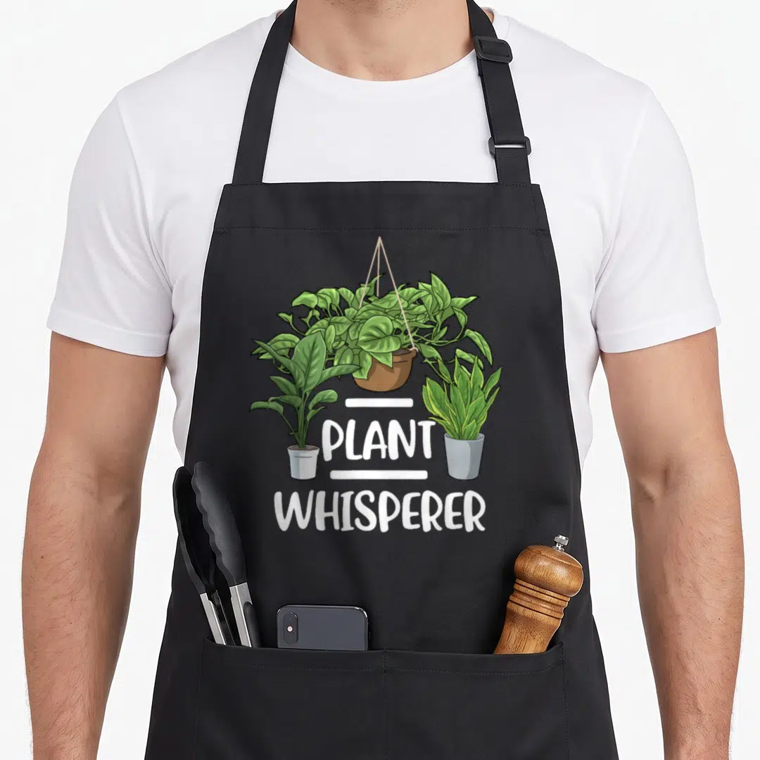 Whisperer gardener apron - Three houseplants including a hanging vine above PLANT WHISPERER text in white cartoon — shown on a black cotton canvas apron against a white background