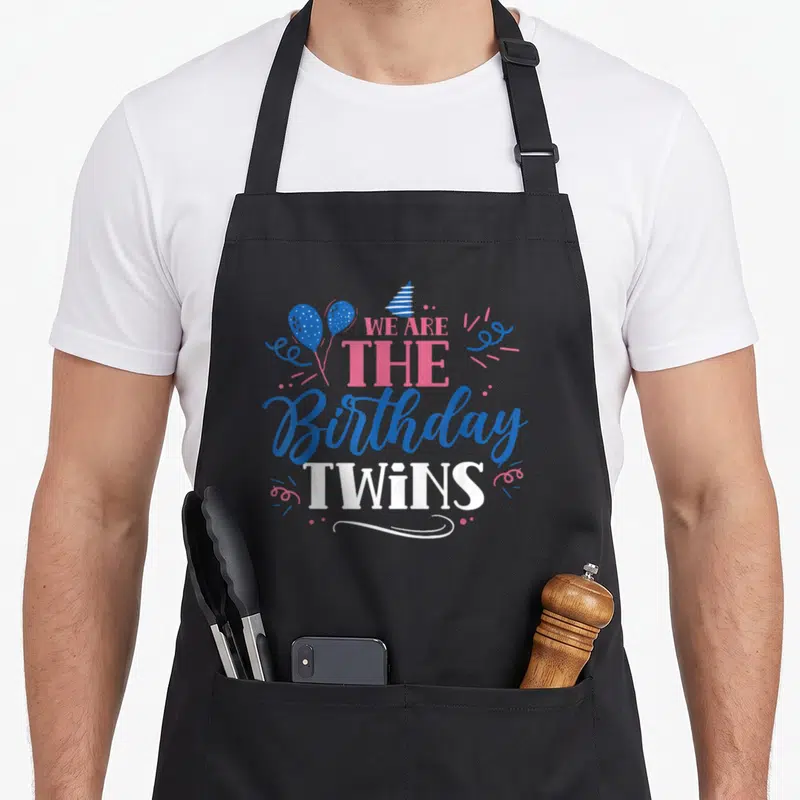 Cool twin birthday apron - Stacked text WE ARE THE Birthday TWINS with blue pink balloons and party hat — shown on a black cotton canvas apron against a white background