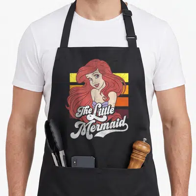 Cute stripe apron - Cartoon mermaid with red hair on red yellow orange and purple stripes with white script text — shown on a black cotton canvas apron against a white background