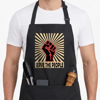 Communist to serve man apron - Raised fist with red outline over beige sunburst and serve the people text — shown on a black cotton canvas apron against a white background