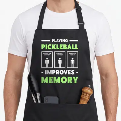Playing pickleball apron - White and lime green text asking what score and who served above three stick figure — shown on a black cotton canvas apron against a white background