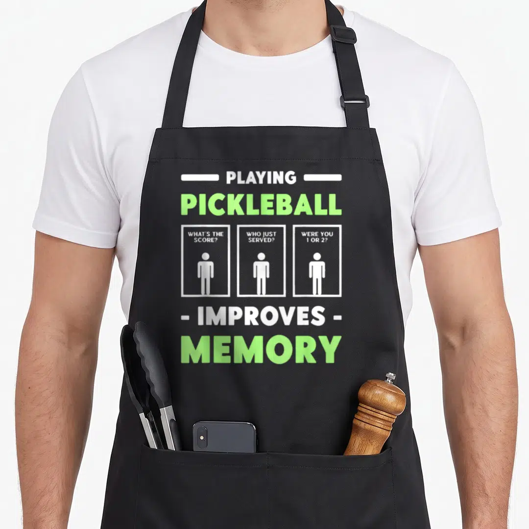 Playing pickleball apron - White and lime green text asking what score and who served above three stick figure — shown on a black cotton canvas apron against a white background