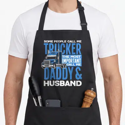 Trucker driving aprons - Semi-truck graphic with blue and white text reading Trucker Daddy Husband — shown on a black cotton canvas apron against a white background