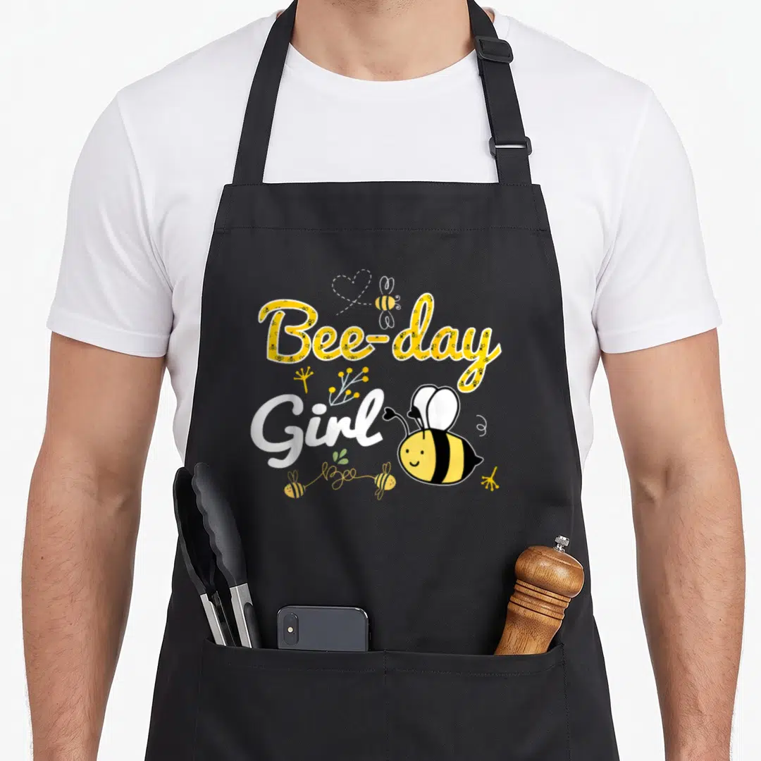 Funny birthday bee aprons - Bee-day Girl text with cartoon bees and flowers in yellow white and black — shown on a black cotton canvas apron against a white background