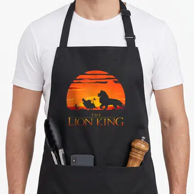 Sunset lion dad apron - THE LION KING text below lion hyena bird silhouettes on orange sun — shown on a black cotton canvas apron against a white background