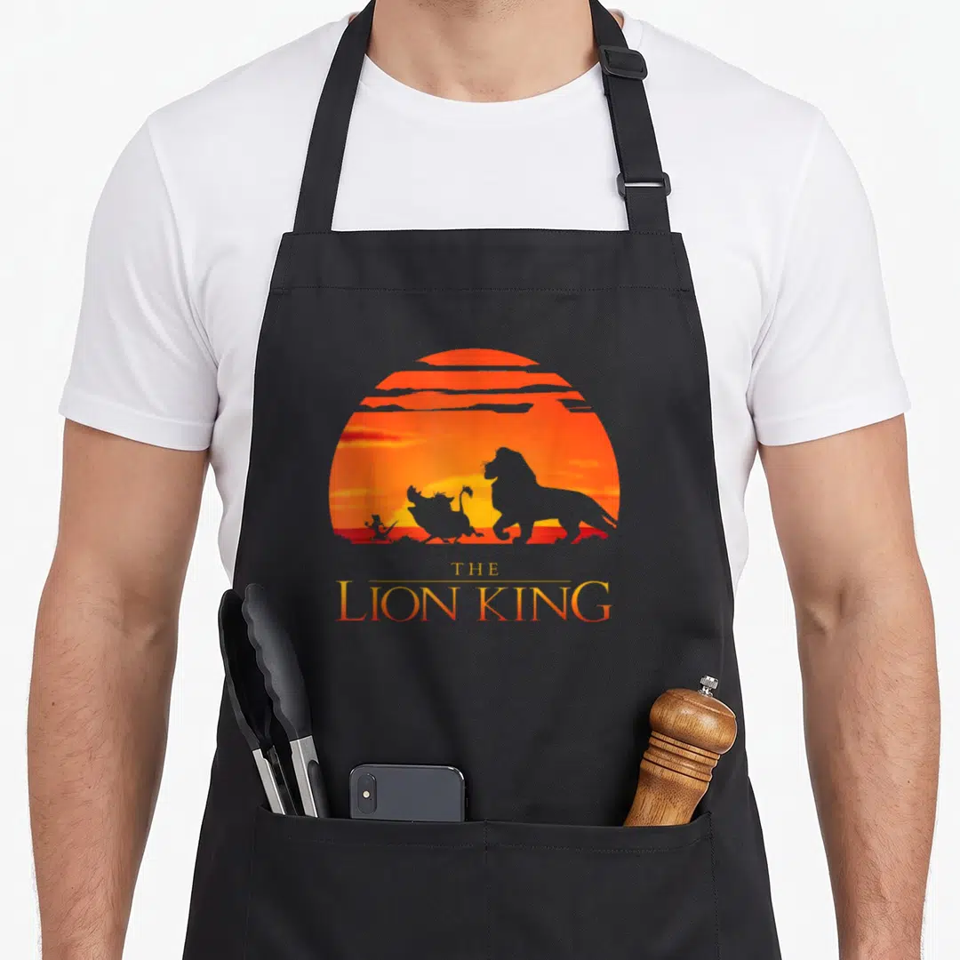 Sunset lion dad apron - THE LION KING text below lion hyena bird silhouettes on orange sun — shown on a black cotton canvas apron against a white background