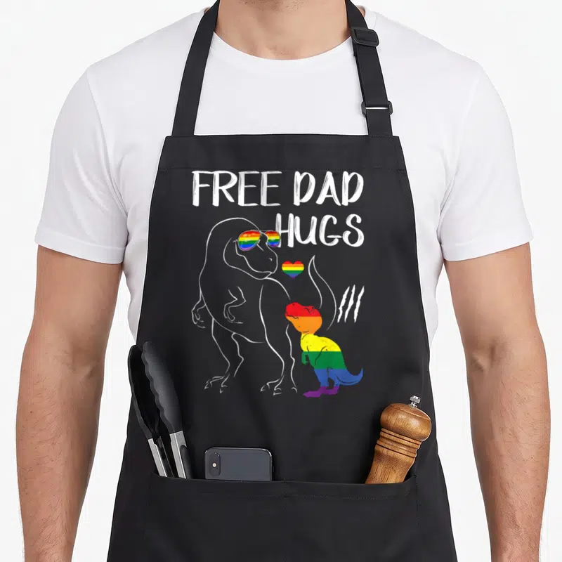 Free dinosaur apron - FREE DAD HUGS text with outlined T-Rex sunglasses and rainbow dino heart — shown on a black cotton canvas apron against a white background