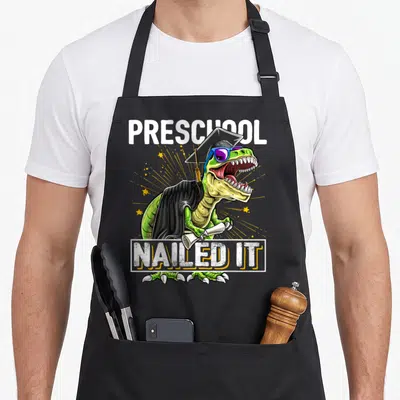 Nailed dinosaur apron - Green dinosaur wearing cap gown sunglasses holding diploma with PRESCHOOL NAILED IT text — shown on a black cotton canvas apron against a white background