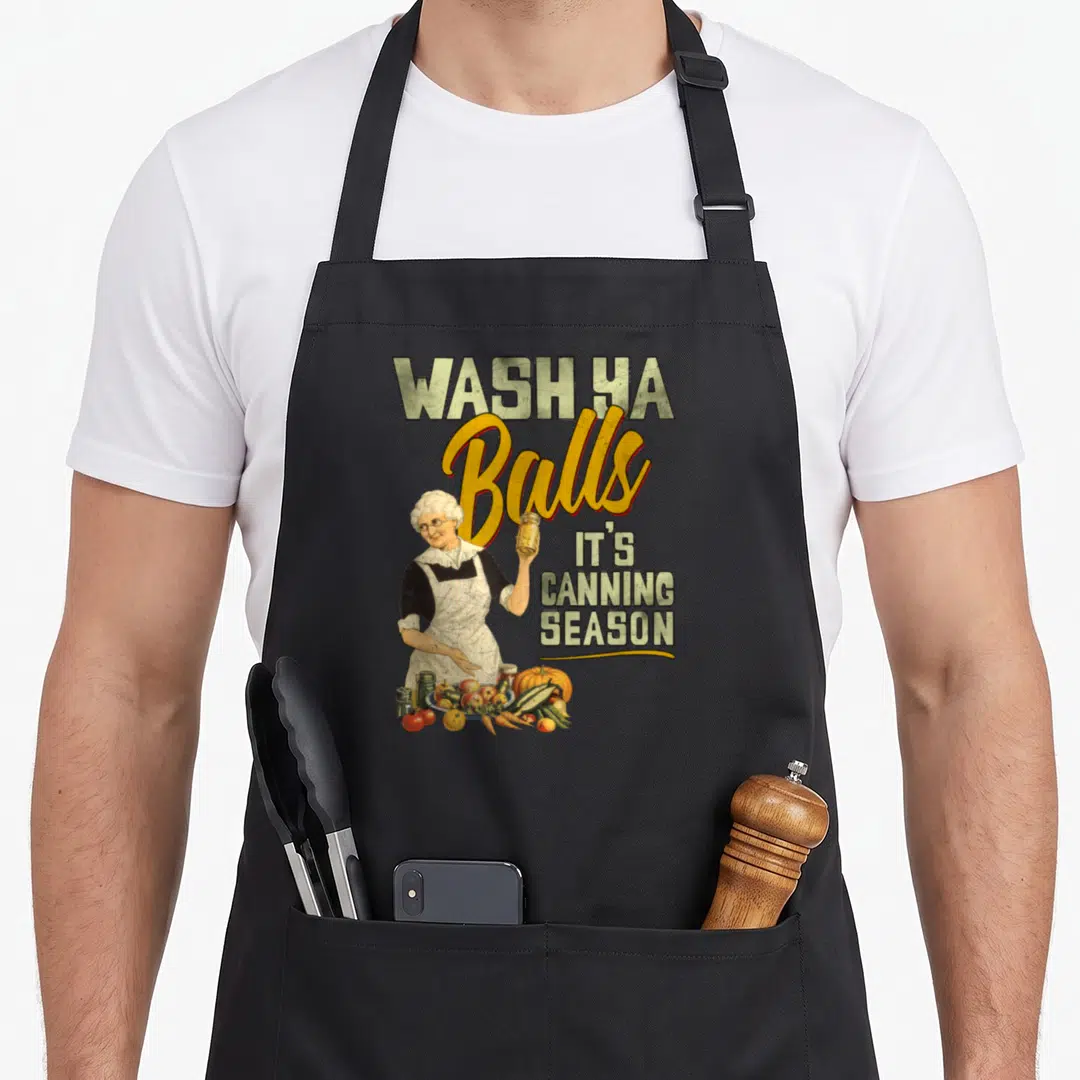 Wash canning apron - Retro illustration of elderly woman holding jar with text WASH YA Balls IT'S CANNING SEASON — shown on a black cotton canvas apron against a white background